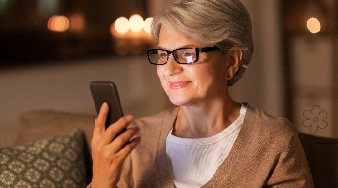 Smartphone auch als Senior vielseitig und sicher nutzen Eine grauhaarige Frau mit Brille sitzt auf dem Sofa. Sie schaut in ihr Smartphone und lächelt.